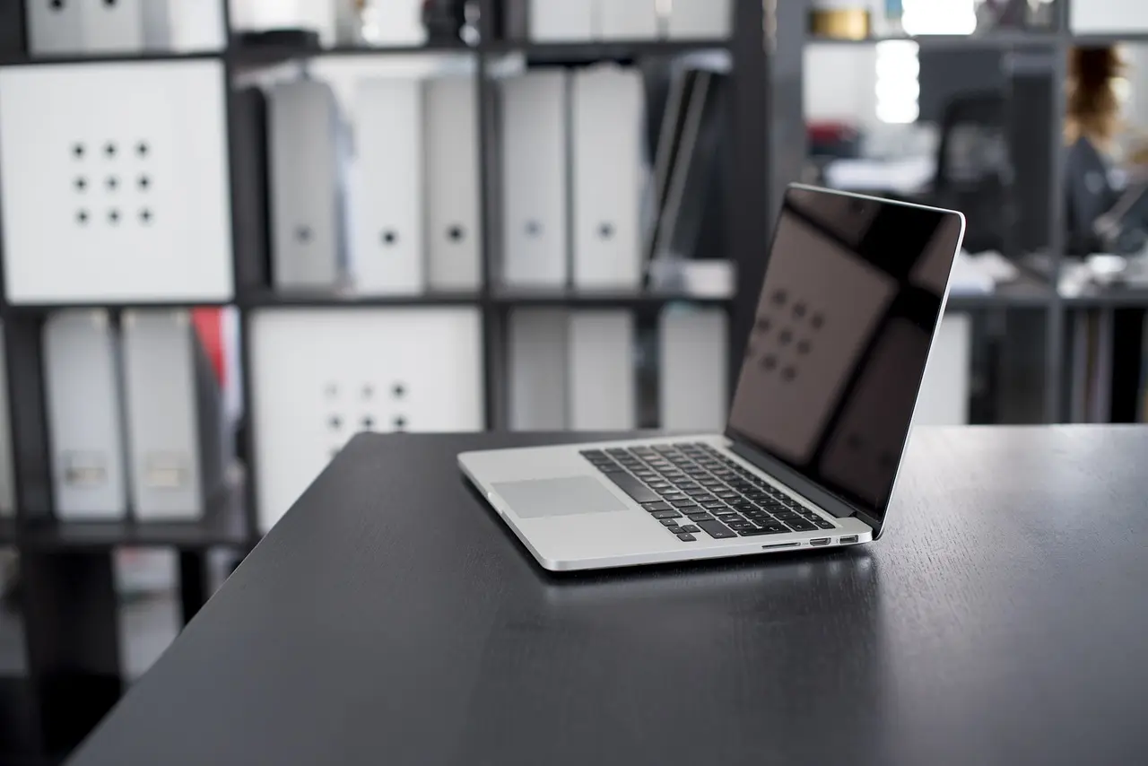 laptop stojący na czarnym biurku, w tle regały z dokumentami